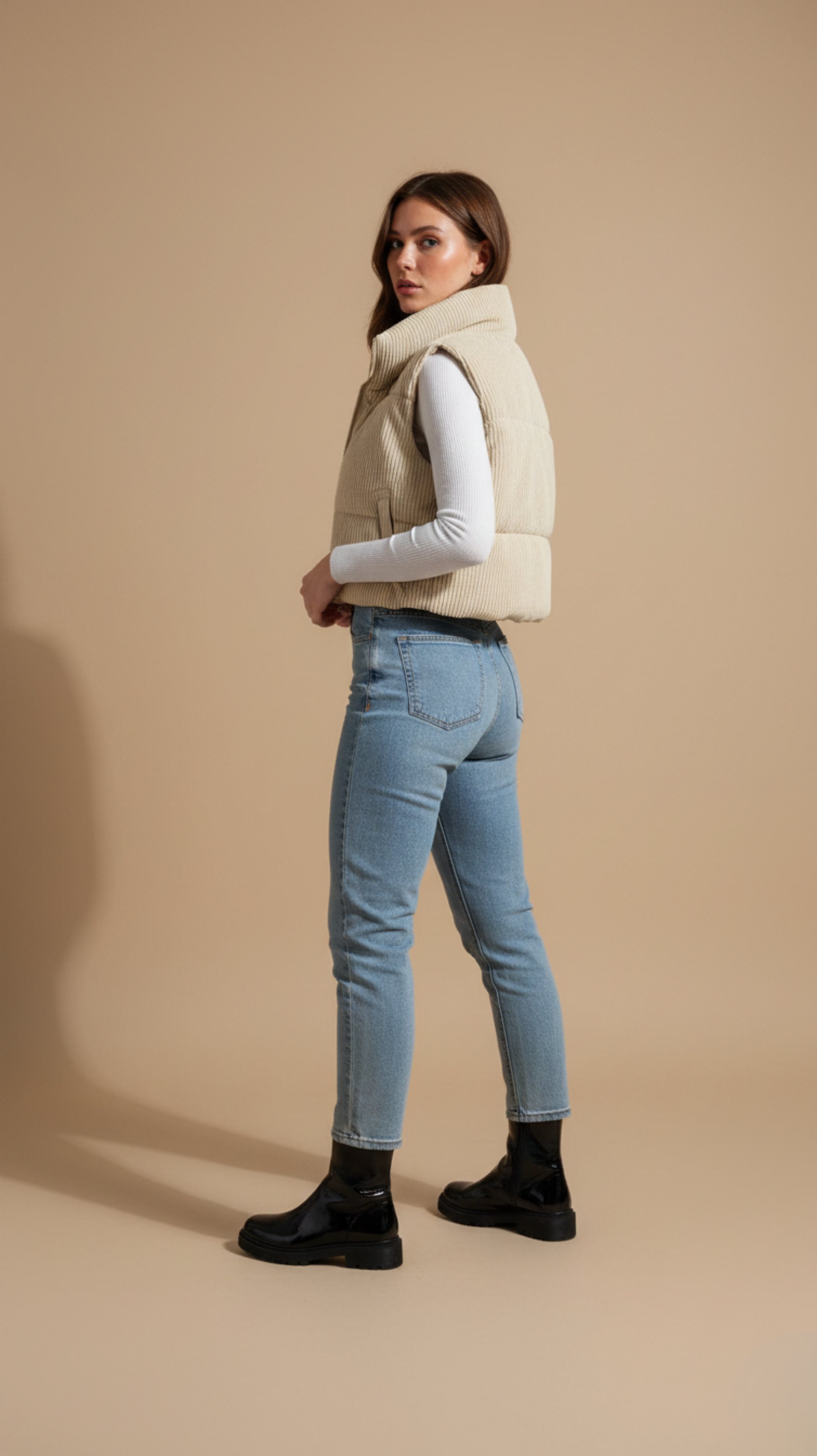 Woman wearing a beige vest, white shirt, blue jeans, and black boots on a beige background