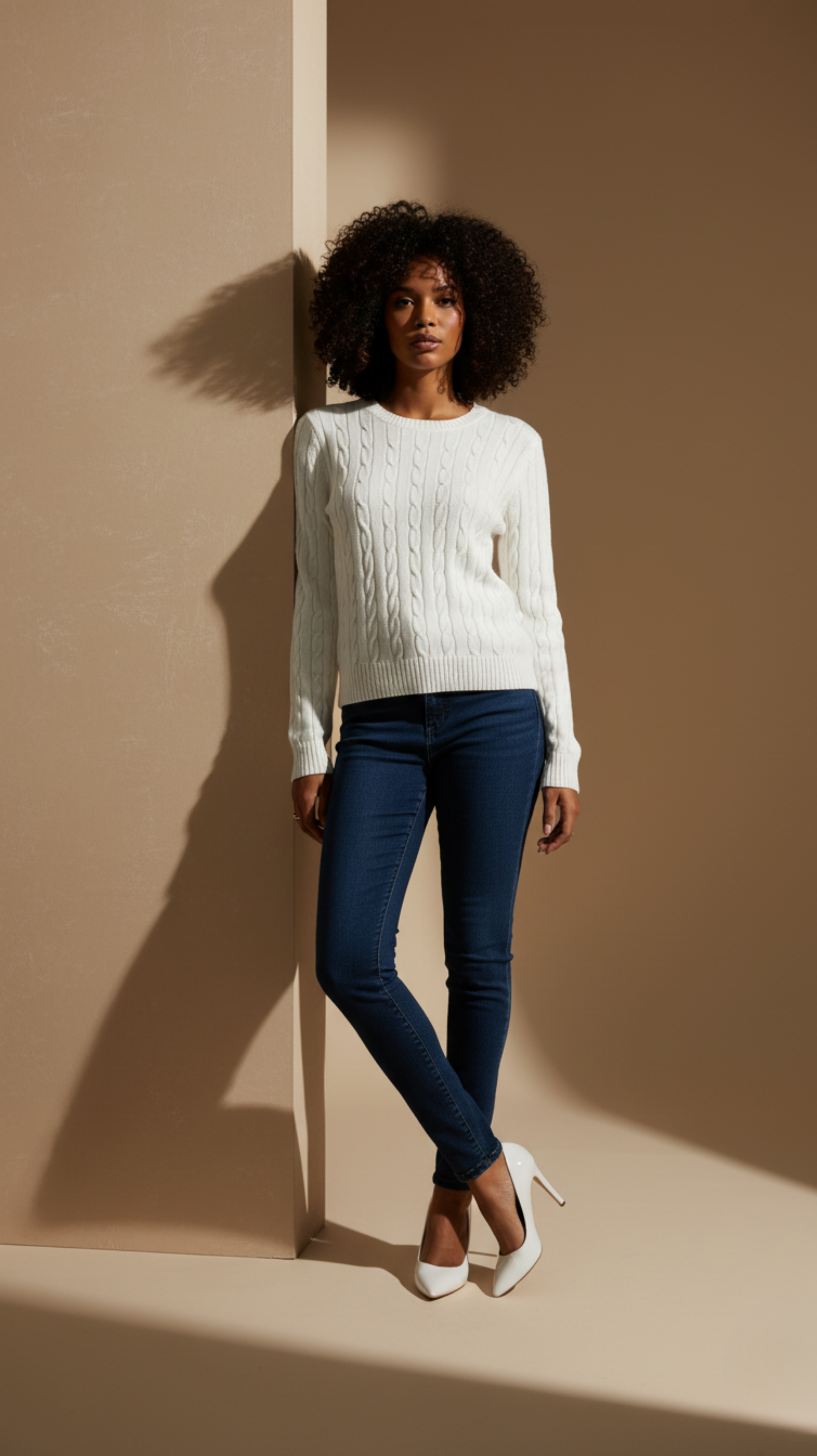 Woman wearing a white sweater and blue jeans standing against a beige wall.